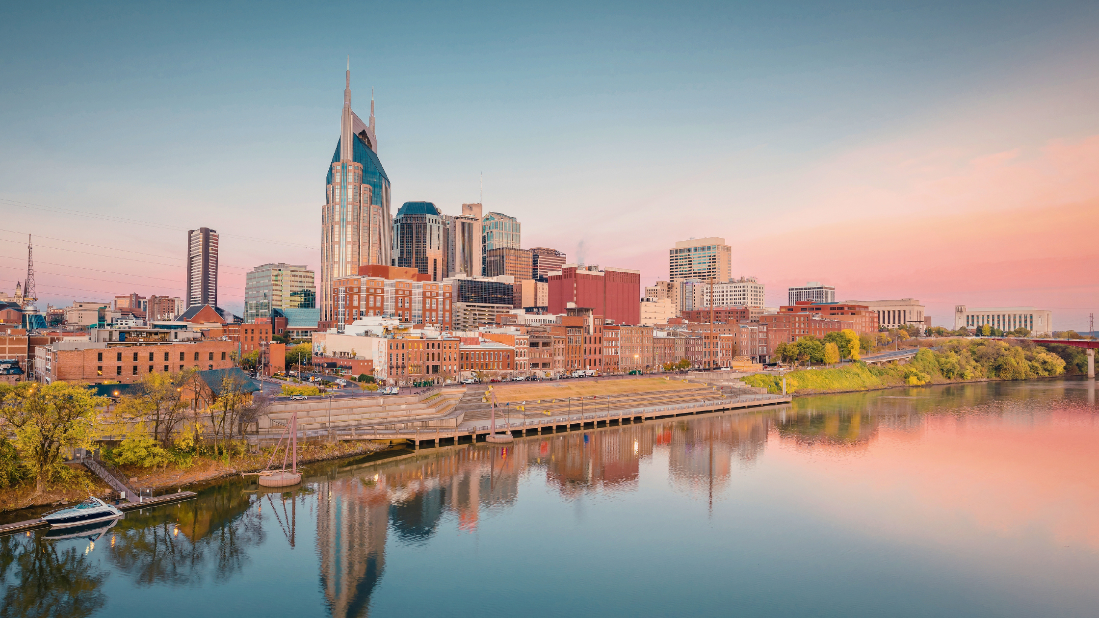 A wide shot of the Nashville skyline at sunset, with the Cumberland River in the foreground reflecting the colorful sky and buildings.