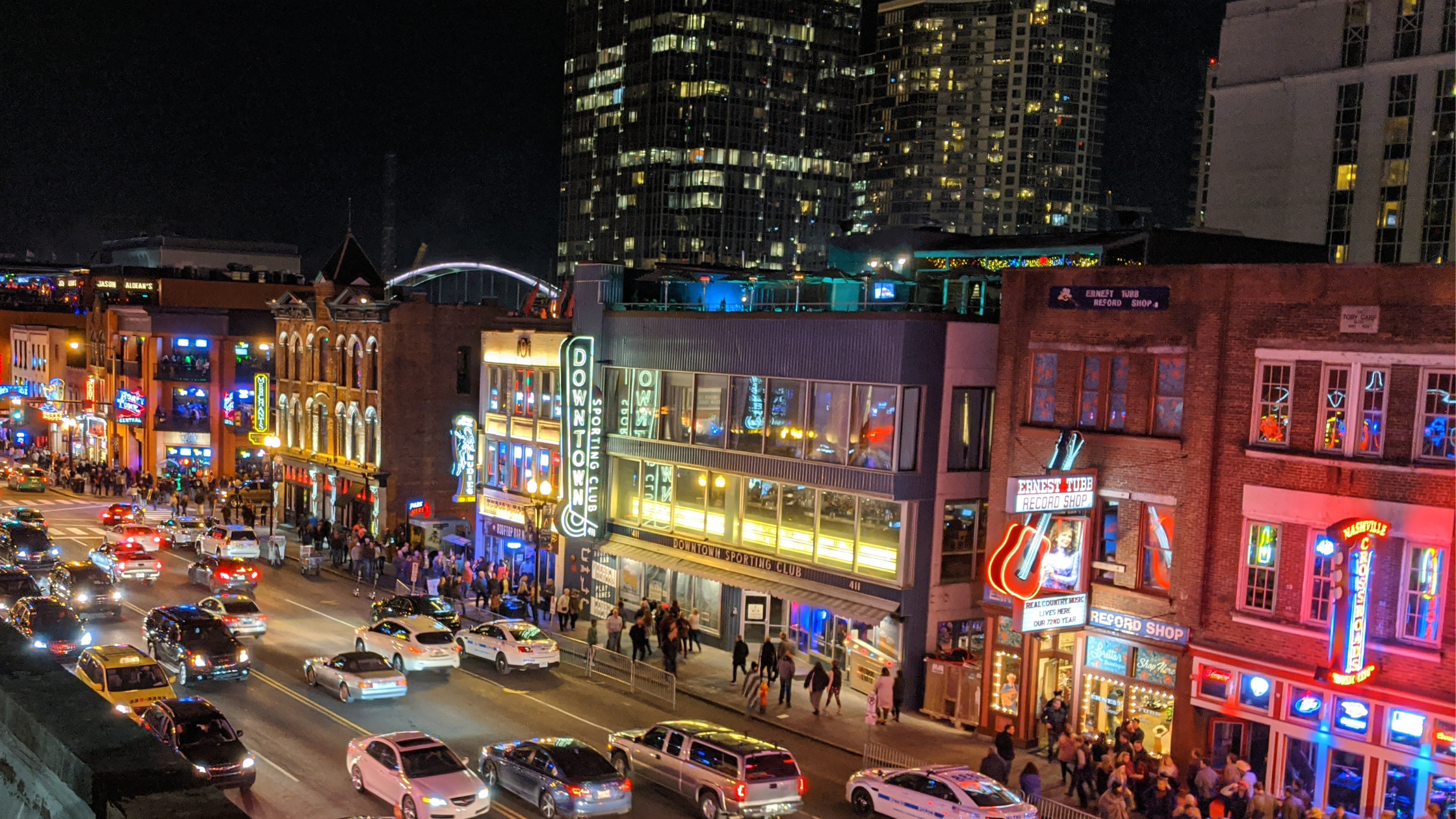 A bustling city street at night, lined with illuminated buildings and neon signs, with cars driving and pedestrians walking on the sidewalks.
